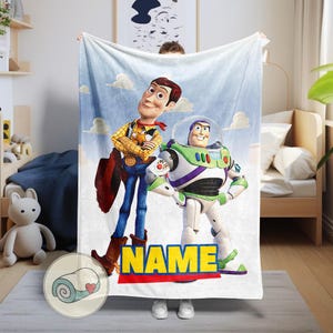 Pu&ograve; includere: Una coperta bianca con Woody e Buzz Lightyear di Toy Story su uno sfondo di cielo blu con nuvole. La coperta ha la parola "NAME" in lettere gialle con un bordo rosso e blu.