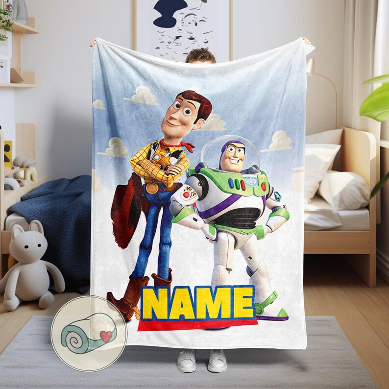 Pu&ograve; includere: Una coperta bianca con Woody e Buzz Lightyear di Toy Story su uno sfondo di cielo blu con nuvole. La coperta ha la parola "NAME" in lettere gialle con un bordo rosso e blu.