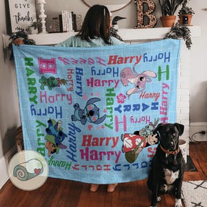 K&ouml;nnte beinhalten: Eine hellblaue Decke mit Cartoon-Figuren und dem Namen "Harry" in verschiedenen Schriftarten und Farben. Die Decke hat ein Gittermuster und enth&auml;lt Illustrationen von Stitch und anderen Charakteren. Eine zusammengerollte Decke mit einem Herz befindet sich in der Ecke.