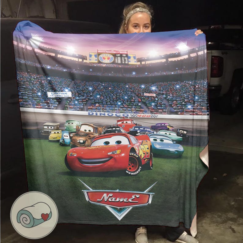 May include: A blanket featuring characters from the animated movie Cars. The design includes Lightning McQueen and other race cars on a green track, with a stadium backdrop. The blanket has a blue border and a personalized name area.