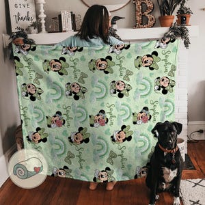 K&ouml;nnte beinhalten: Eine hellgr&uuml;ne Decke mit einem Muster von Minnie Mouse, Schleifen, Regenbogen und dem Namen "Sophia". Die Decke wird von einer Person gehalten. Ein schwarzer Hund sitzt in der N&auml;he. Ein gerolltes Bild befindet sich in der Ecke.