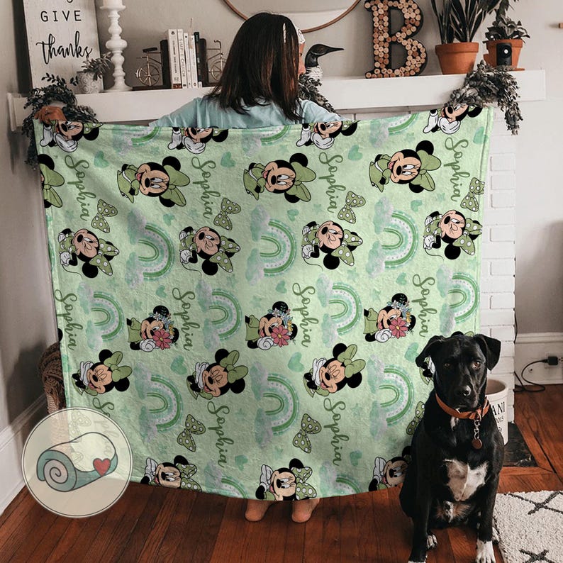 K&ouml;nnte beinhalten: Eine hellgr&uuml;ne Decke mit einem Muster von Minnie Mouse, Schleifen, Regenbogen und dem Namen "Sophia". Die Decke wird von einer Person gehalten. Ein schwarzer Hund sitzt in der N&auml;he. Ein gerolltes Bild befindet sich in der Ecke.