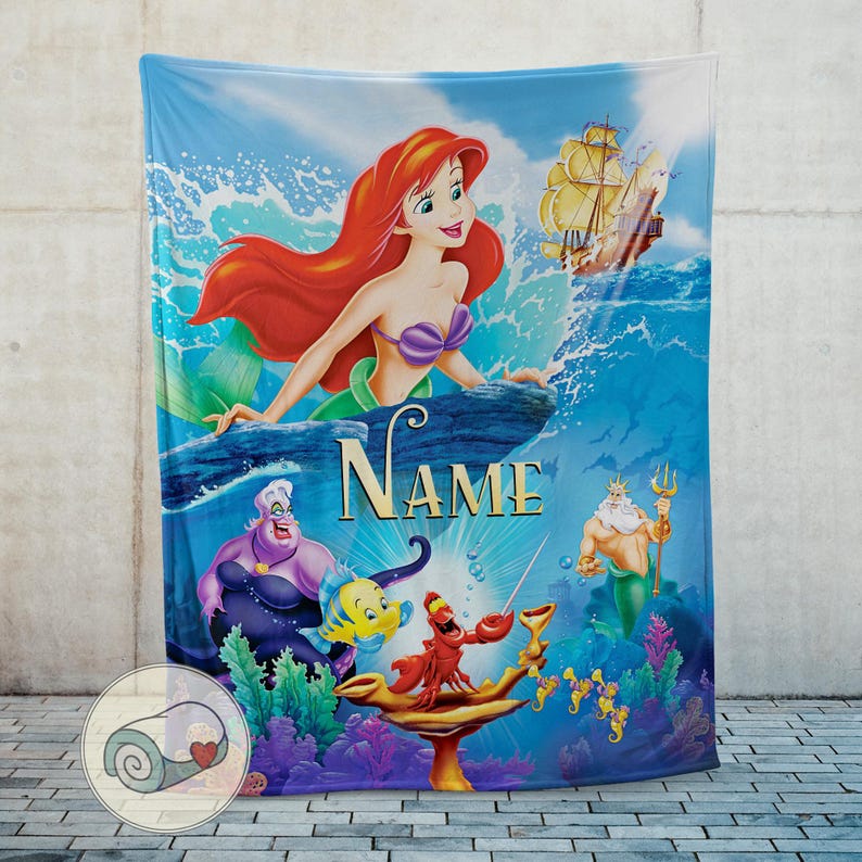 May include: A blue blanket featuring a colorful illustration of Ariel from The Little Mermaid. The design includes Ariel, Ursula, Flounder, Sebastian, King Triton, and a ship. The word "NAME" is printed in gold.
