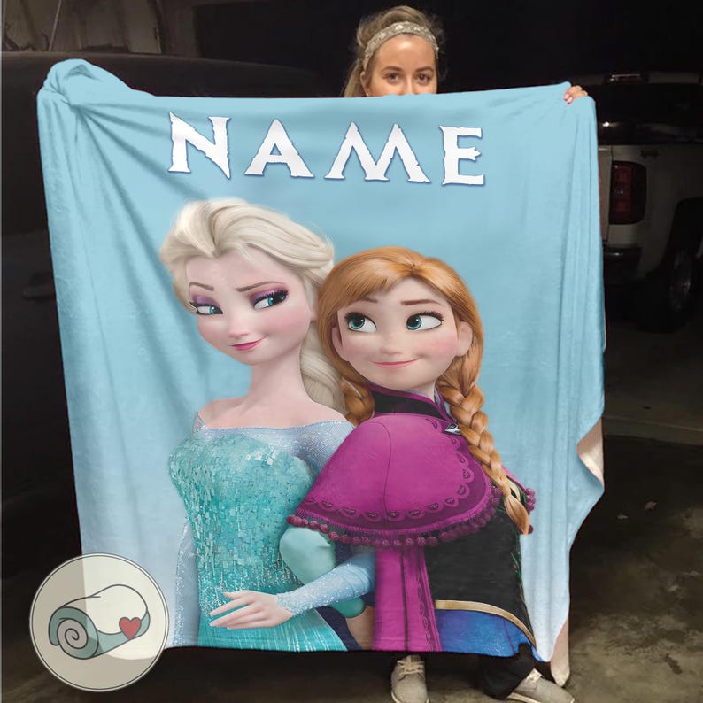 Puede incluir: Manta azul claro con Elsa y Anna de Frozen, con la palabra "NAME" en letras blancas en la parte superior. Elsa lleva un vestido turquesa y Anna una capa morada. Un logotipo de manta enrollada est&aacute; en la esquina inferior izquierda.