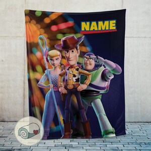 Puede incluir: Una manta de felpa con personajes de la franquicia Toy Story. La manta muestra a Woody, Buzz Lightyear y Bo Peep sobre un fondo azul oscuro con la palabra "NAME" en amarillo. La manta est&aacute; dise&ntilde;ada con colores vibrantes y una textura suave.