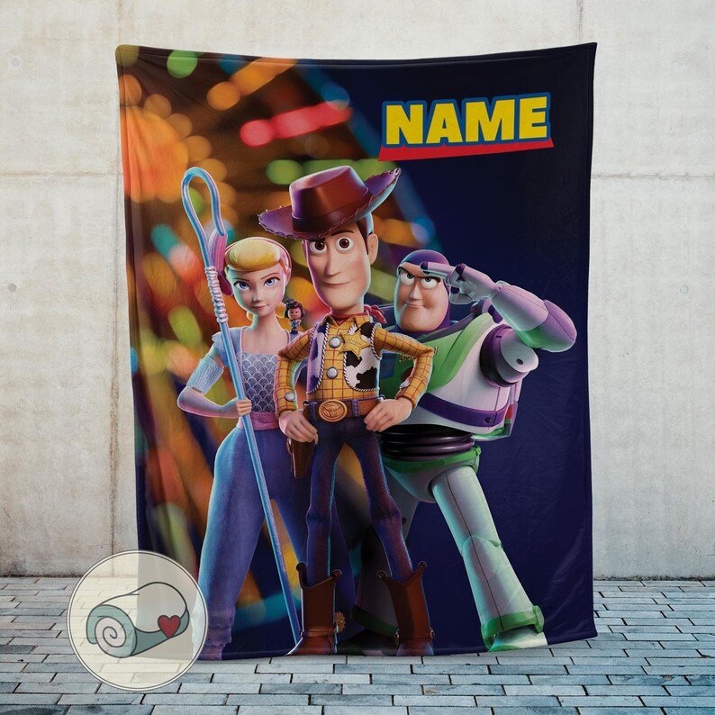 Puede incluir: Una manta de felpa con personajes de la franquicia Toy Story. La manta muestra a Woody, Buzz Lightyear y Bo Peep sobre un fondo azul oscuro con la palabra "NAME" en amarillo. La manta est&aacute; dise&ntilde;ada con colores vibrantes y una textura suave.
