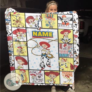 Op de afbeelding: Een kleurrijke deken met illustraties van Jessie uit Toy Story. De deken heeft een witte achtergrond met zwarte koeienvlekken op de rand. Het woord "NAME" wordt weergegeven in een blauw en geel kader, met een lasso-afbeelding.