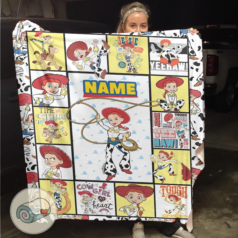 Op de afbeelding: Een kleurrijke deken met illustraties van Jessie uit Toy Story. De deken heeft een witte achtergrond met zwarte koeienvlekken op de rand. Het woord "NAME" wordt weergegeven in een blauw en geel kader, met een lasso-afbeelding.
