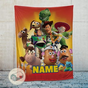 May include: A red and yellow blanket featuring a group of characters from the Toy Story movie franchise. The blanket has a red border and the word "NAME" in yellow letters.