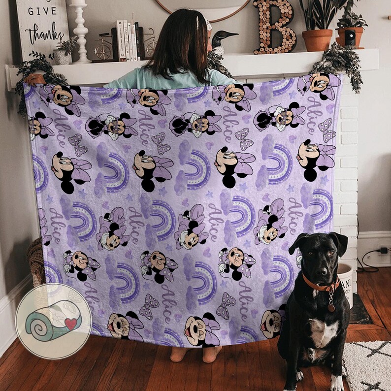 Op de afbeelding: Een lavendelkleurige deken met een herhaald patroon van Minnie Mouse, regenbogen en de naam "Alice". De deken heeft een zachte, pluche uitstraling, geschikt voor een kinderkamer of als decoratief kleed.
