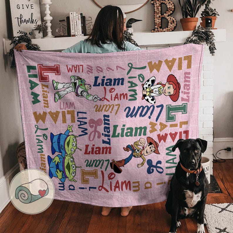 May include: A pink blanket featuring cartoon characters and the name "Liam" in various fonts and colors. The blanket includes images of a space ranger, a cowgirl, and other characters. A black dog sits in front of the blanket.