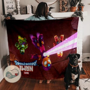 Op de afbeelding: Een bordeauxrode deken met kleurrijke stripfiguren en de tekst "NAME AWESOME FRIENDS" met het Marvel-logo. De deken heeft een opgerold beeld in de hoek. Een zwarte hond met een bruine halsband zit voor de deken.