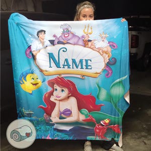 May include: A blue blanket featuring Ariel from The Little Mermaid, with Flounder, Sebastian, Prince Eric, Ursula, and King Triton. The blanket has a personalized name banner and a rolled-up blanket icon.