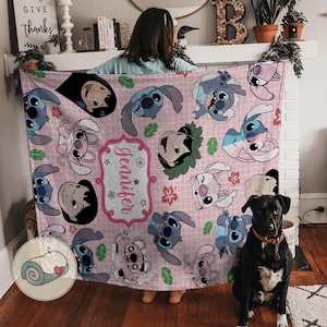 May include: A pink blanket featuring cartoon characters and the name "Jennifer" in a decorative frame. The blanket has a repeating pattern of the characters and floral accents. The blanket is being held up by a person.