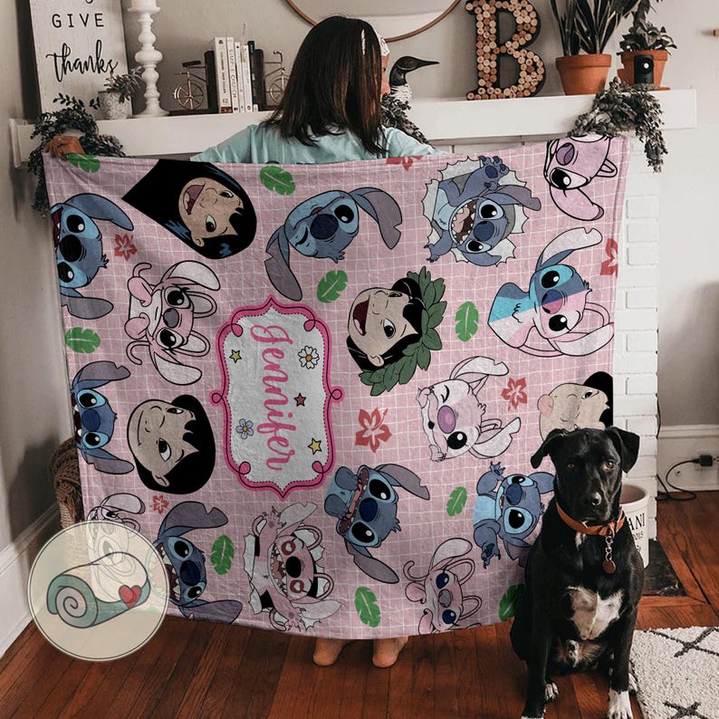 May include: A pink blanket featuring cartoon characters and the name "Jennifer" in a decorative frame. The blanket has a repeating pattern of the characters and floral accents. The blanket is being held up by a person.