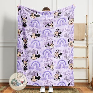 Op de afbeelding: Een lavendelkleurige deken met een herhalend patroon van Minnie Mouse, regenbogen, strikken en de naam "Alice". Het ontwerp bevat verschillende poses van Minnie Mouse, wolken en sterren. De deken wordt door een persoon vastgehouden.