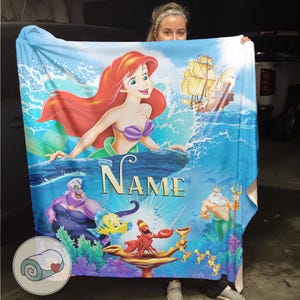 May include: A blue blanket featuring Ariel from The Little Mermaid. The blanket has a vibrant illustration of Ariel, a ship, and other characters from the movie. The word "NAME" is printed on the blanket.