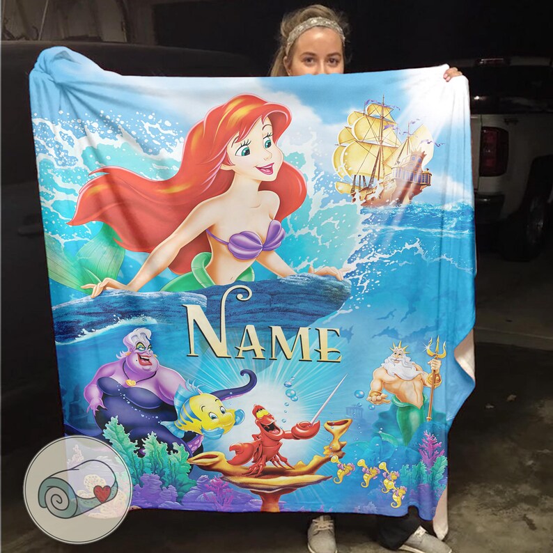 May include: A blue blanket featuring Ariel from The Little Mermaid. The blanket has a vibrant illustration of Ariel, a ship, and other characters from the movie. The word "NAME" is printed on the blanket.