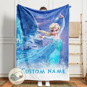 Puede incluir: Una manta polar azul con un gr&aacute;fico de una mujer rubia con un vestido azul, copos de nieve y un fondo helado. El texto "CUSTOM NAME" est&aacute; impreso en azul en la parte inferior. La manta est&aacute; sujeta por una persona.