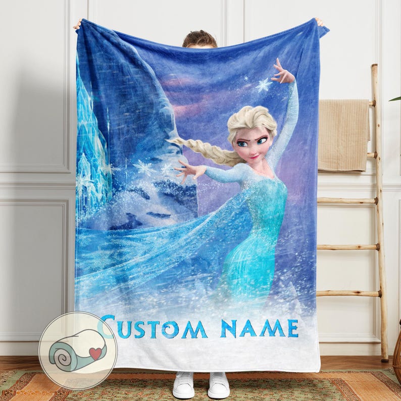 Puede incluir: Una manta polar azul con un gr&aacute;fico de una mujer rubia con un vestido azul, copos de nieve y un fondo helado. El texto "CUSTOM NAME" est&aacute; impreso en azul en la parte inferior. La manta est&aacute; sujeta por una persona.