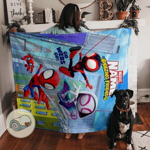 May include: A blue blanket featuring Spider-Man and other characters from the Marvel universe. The blanket has a colorful design with the words "Amazing Friends" and "Go Webs Go!" printed on it. A black dog sits in front of the blanket.