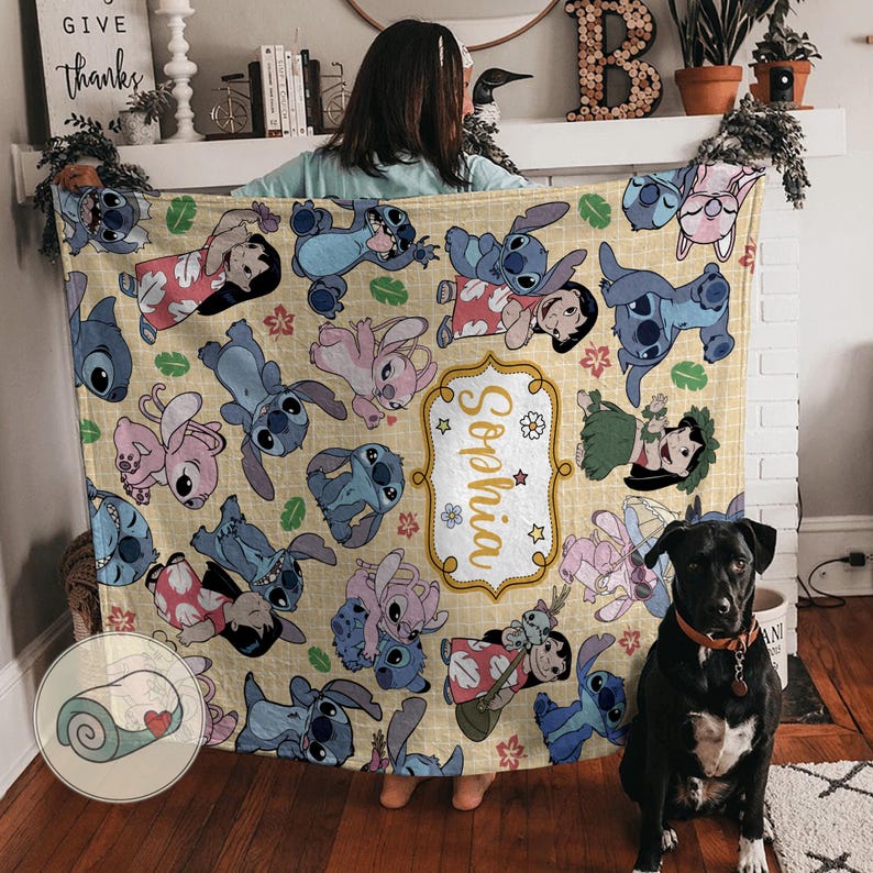 Pu&ograve; includere: Una coperta beige con un motivo ripetuto di personaggi dei cartoni animati blu, rosa e rossi. La coperta ha un'etichetta personalizzata con il nome "Sophia" in oro. La coperta &egrave; piegata e tenuta da una persona.