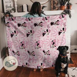 K&ouml;nnte beinhalten: Eine rosa Decke mit einem sich wiederholenden Muster von Minnie Mouse, Schleifen, Regenbogen und dem Namen "Alice". Die Decke hat ein aufgerolltes Bild in der Ecke. Die Decke wird von einer Person gehalten.