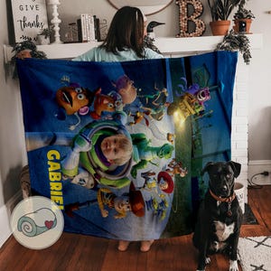 May include: A blue blanket featuring characters from the Toy Story movie, including Buzz Lightyear and Woody. The blanket has the name "Gabriel" printed on it. A black dog with a brown collar sits in front of the blanket.
