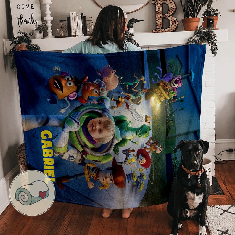 May include: A blue blanket featuring characters from the Toy Story movie, including Buzz Lightyear and Woody. The blanket has the name "Gabriel" printed on it. A black dog with a brown collar sits in front of the blanket.