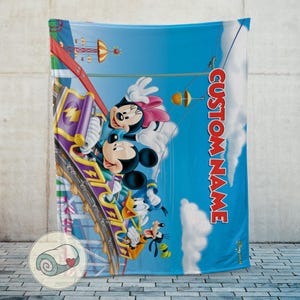 May include: A blue blanket featuring Mickey Mouse, Minnie Mouse, Donald Duck, and Goofy on a roller coaster. The blanket has the text "CUSTOM NAME" in red. The background is a blue sky with amusement park elements.