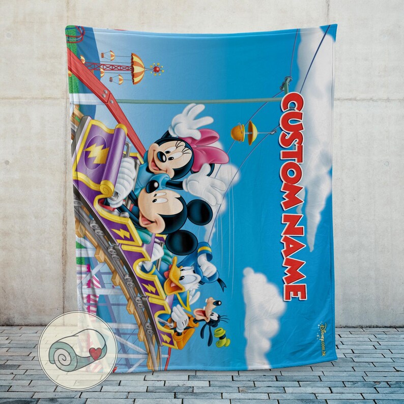 May include: A blue blanket featuring Mickey Mouse, Minnie Mouse, Donald Duck, and Goofy on a roller coaster. The blanket has the text "CUSTOM NAME" in red. The background is a blue sky with amusement park elements.