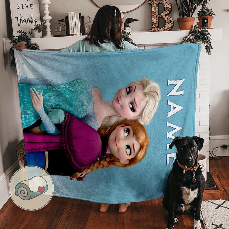 Puede incluir: Una manta azul claro con Elsa y Anna de Frozen, con el nombre "NAME" impreso verticalmente en blanco. Un perro negro con collar marr&oacute;n est&aacute; cerca.