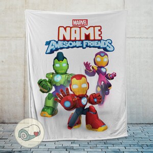 Op de afbeelding: Witte deken met drie striphelden: Iron Man, Hulk en een ander personage. De deken heeft het Marvel-logo en de woorden "NAME" en "Awesome Friends" in een kleurrijk lettertype. De deken wordt getoond tegen een lichtgrijze muur.