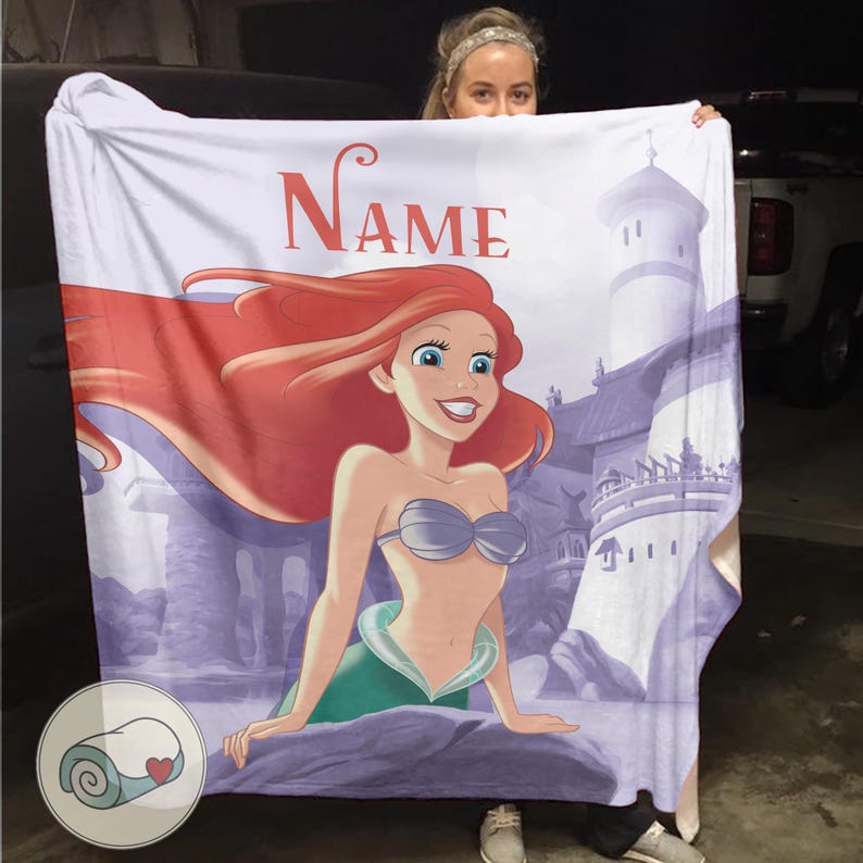 May include: A white blanket featuring a cartoon illustration of a mermaid with red hair, a silver seashell bra, and a green tail. The word "Name" is printed in red above the mermaid. A castle is in the background.