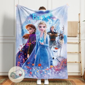 May include: A large, rectangular blanket featuring characters from the movie "Frozen." The blanket displays Elsa, Anna, Kristoff, Olaf, and Sven against a snowy mountain backdrop. The word "ELSA" is at the top in blue letters. The blanket is held up by a person.
