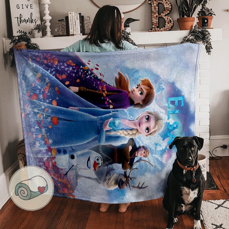 May include: A large, rectangular fleece blanket featuring characters from the animated film "Frozen." The blanket displays Anna, Elsa, Kristoff, Olaf, and Sven against a snowy, blue background. The word "Elsa" is written in blue script.