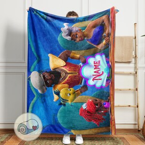 May include: A blue fleece blanket with cartoon characters from a popular animated film. The blanket has a repeating pattern and a personalised name graphic. The characters include a mermaid, a sea witch, and a prince.