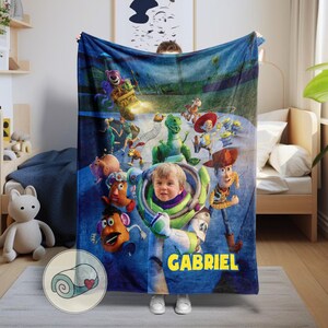 May include: A blue blanket featuring characters from the animated film Toy Story, including Buzz Lightyear, Woody, and Rex. The blanket has a personalized name, "GABRIEL," in yellow block letters. The blanket is made of soft material.