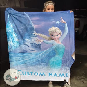 Puede incluir: Una manta azul con una imagen de dibujos animados de una mujer con cabello rubio con un vestido azul, con el texto "CUSTOM NAME" en la parte inferior. El dise&ntilde;o incluye copos de nieve y un castillo helado.