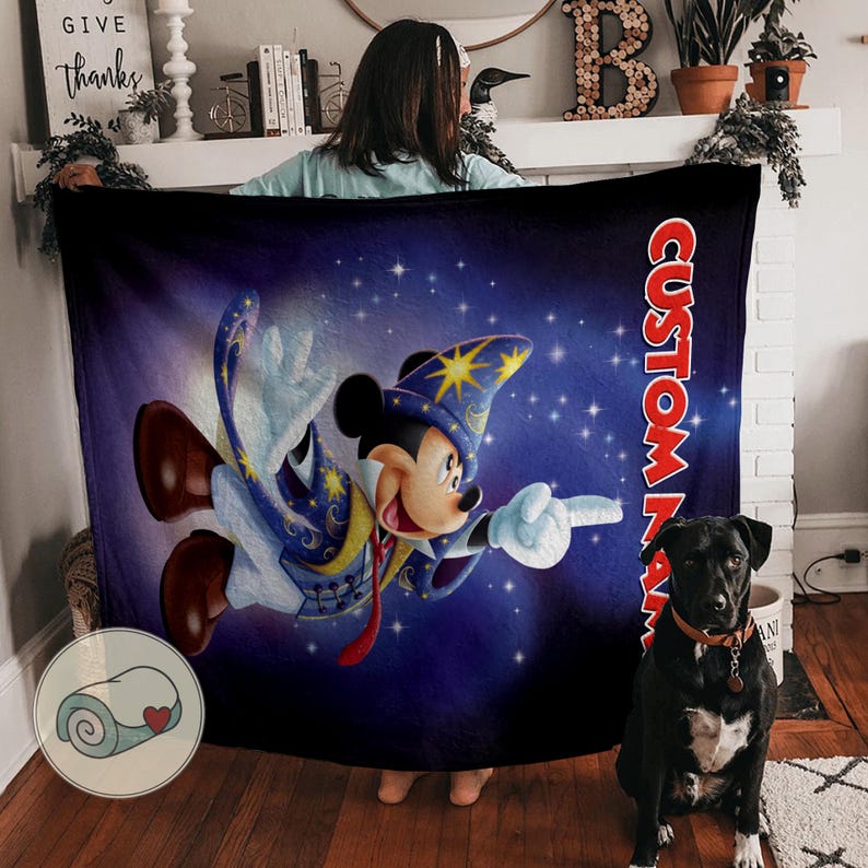 Op de afbeelding: Een marineblauwe deken met een cartoonafbeelding van Mickey Mouse als tovenaar, tegen een sterrenhemel. De deken heeft de tekst "CUSTOM NAME" in het rood. De deken is opgerold in een kleine cirkel.