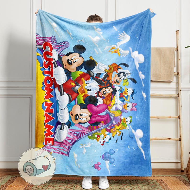 May include: A large, blue blanket featuring a colorful illustration of Mickey Mouse, Minnie Mouse, Donald Duck, and other Disney characters. The blanket has a yellow stripe with the text "CUSTOM NAME" and a roller coaster design. A rolled-up blanket is shown in a small circle.