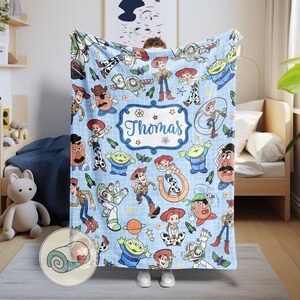May include: A light blue blanket featuring cartoon characters from the Toy Story franchise. The blanket has a repeating pattern of Woody, Buzz Lightyear, Jessie, and other characters. The name "Thomas" is displayed in a decorative frame.