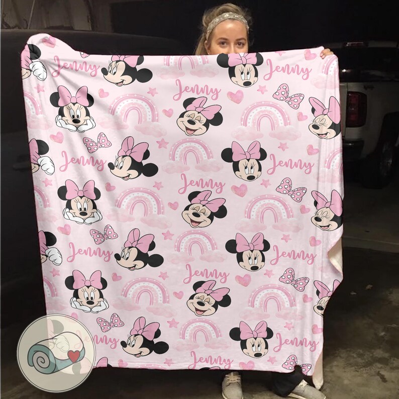 May include: A pink blanket featuring Minnie Mouse, rainbows, hearts, and bows. The blanket has the name "Jenny" printed across it in a cursive font. The design is repeated across the entire blanket.