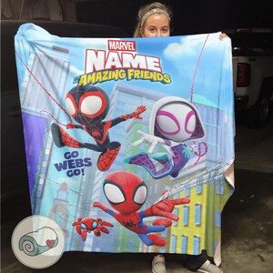 May include: A Marvel "Amazing Friends" blanket featuring cartoon Spider-Man characters. The blanket has a blue background with city buildings and the words "Go Webs Go!". The blanket is held up by a person.