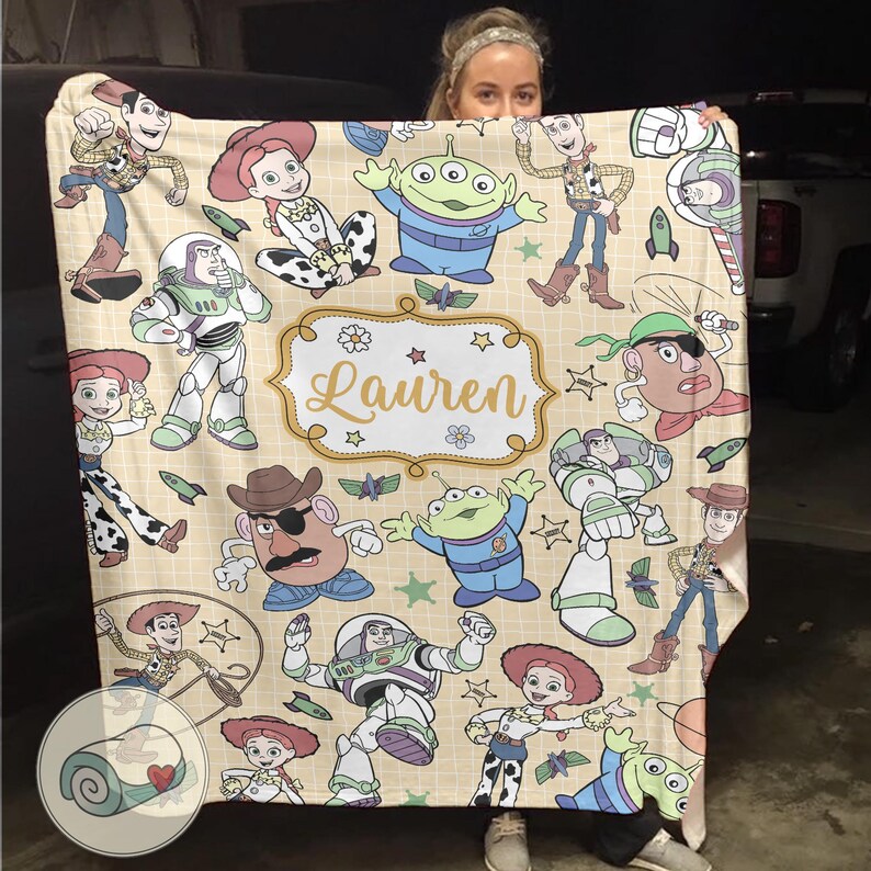 May include: A beige blanket featuring cartoon characters from the Toy Story franchise. The blanket has a repeating pattern of Woody, Buzz Lightyear, Jessie, and other characters. The name "Lauren" is written in a decorative frame.
