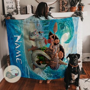 May include: A large, rectangular blue blanket featuring a vibrant illustration of characters from a popular animated film. The blanket has a wave design and the word "NAME" printed vertically. A black dog sits in front of the blanket.
