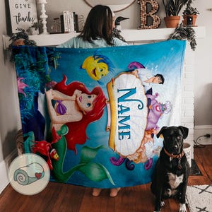May include: A blue fleece blanket featuring characters from The Little Mermaid. The blanket has a central scroll with the word "NAME" and illustrations of Ariel, Flounder, Sebastian, Prince Eric, Ursula, and other characters.