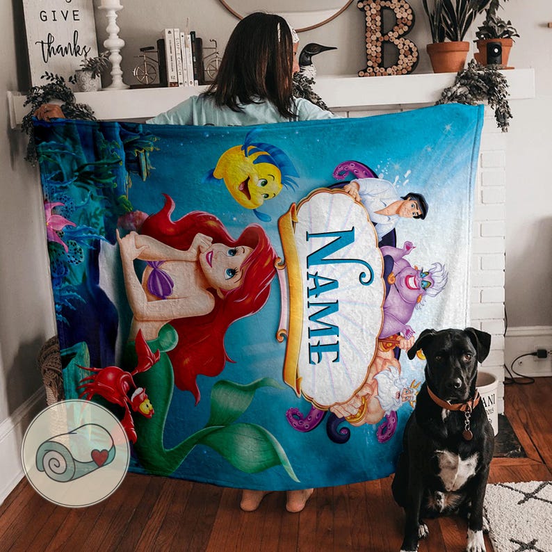 May include: A blue fleece blanket featuring characters from The Little Mermaid. The blanket has a central scroll with the word "NAME" and illustrations of Ariel, Flounder, Sebastian, Prince Eric, Ursula, and other characters.
