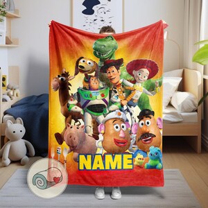 May include: A red and yellow blanket featuring a colorful graphic of characters from the animated film Toy Story. The blanket has the word "NAME" in yellow block letters at the bottom.
