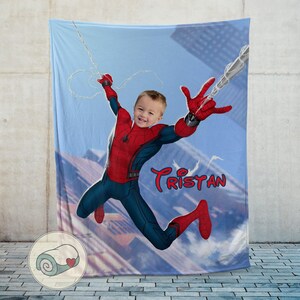 Op de afbeelding: Een gepersonaliseerde Spider-Man-deken met een kind in een Spider-Man-kostuum tegen een stadsachtergrond. De deken is voornamelijk blauw, rood en marineblauw, met de naam "Tristan" in rood script.